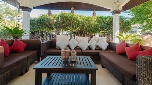 An outdoor seating area with wicker furniture and cushions, a table with decorative items, and a backdrop of greenery and architectural columns. - Kesorn Charming Authentic Thai Pool Villa Phuket (Rawai)