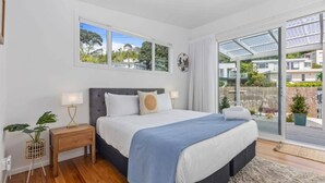 A bedroom with a large bed, a nightstand, a lamp, and a view of the outside through a glass door. - Escape to Kohi Cottage - Your Beachside Haven (Auckland)