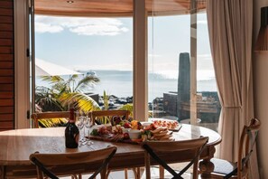 A round table with a bottle of wine, glasses, and a variety of appetizers. The table is set against a large window overlooking a coastal view with palm trees and a cityscape. - Takapuna Beachfront w/Stunning Views (Auckland)