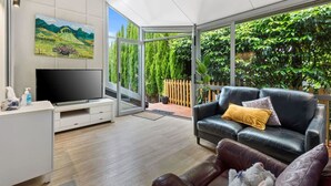 A modern living room with a TV, sofa, and a large window offering a view of greenery. - Ocean View Spacious 5BR Retreat for Families (Auckland)