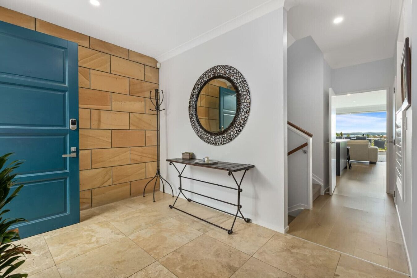 A hallway with a blue door, a wooden wall, a metal console table, a decorative mirror, and a view to a balcony.