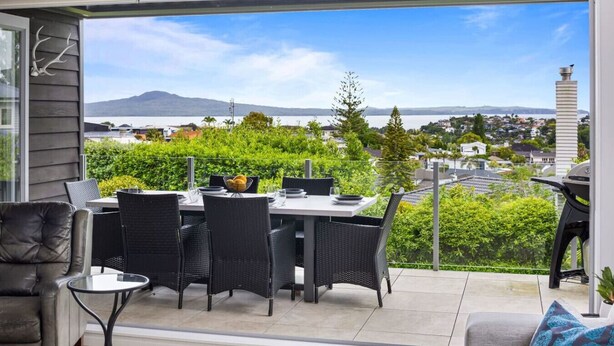 A balcony with a dining table and chairs, a sofa, and a view of a cityscape and mountains. - Ocean View Spacious 5BR Retreat for Families (Auckland)