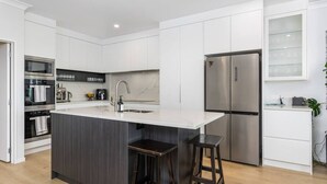 A modern kitchen with white cabinetry, a central island with stools, stainless steel appliances, and a built-in oven. - Ocean View Spacious 5BR Retreat for Families (Auckland)