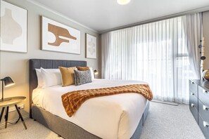 A modern hotel room with a large bed, a bedside table with a lamp, a dresser, and a large window with sheer curtains. - Luxurious Harbour View Apartment in Downtown (Auckland)