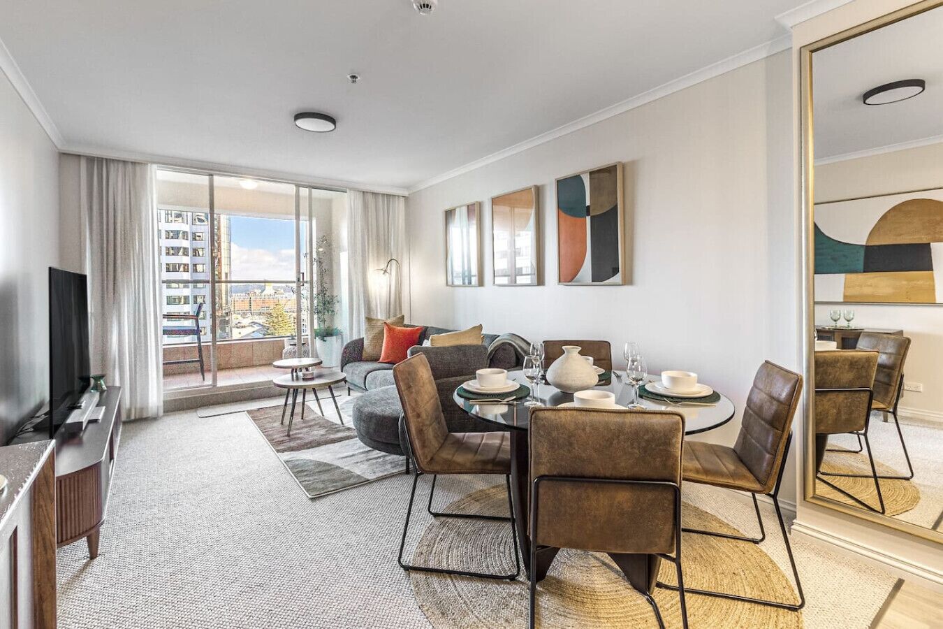 A modern hotel room with a balcony, a dining area with a round table and chairs, and a large mirror reflecting the room.