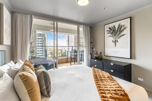 A modern bedroom with a large bed, a view of the city through floor-to-ceiling windows, a dark dresser, and a framed palm tree artwork on the wall.