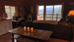 Een gezellig chaletinterieur met een houten eettafel, bankjes, een bank en uitzicht op bergen door grote ramen.