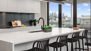 A modern kitchen with a large island, black bar stools, and a city view through large windows. - Stay Devonport - 3 Bedroom Penthouse (Auckland)