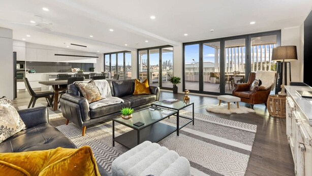 A modern living room with a sofa, coffee table, and a view of the city through large windows. - Stay Devonport - 3 Bedroom Penthouse (Auckland)