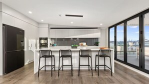A modern kitchen with a central island, bar stools, and a large window offering a city view. - Stay Devonport - 3 Bedroom Penthouse (Auckland)