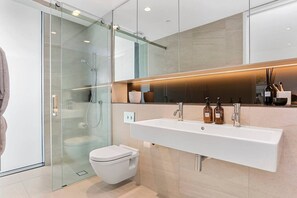 A modern bathroom with a glass shower enclosure, a white toilet, and a large mirror above a white sink with two faucets. - Harbour front Living at Wynyard Marina (Auckland)