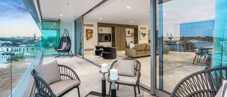 A spacious balcony with a view of the city, furnished with wicker chairs and a table, and a glass wall leading to an interior living area with a sofa and a TV.