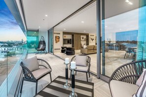 Ein geräumiger Balkon mit Stadtblick, ausgestattet mit Korbstühlen und einem Tisch, und eine Glaswand, die zu einem Wohnbereich mit einer Couch und einem Fernseher führt.