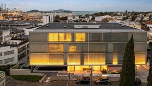 A modern building with a reflective facade, surrounded by residential buildings and a port in the background.