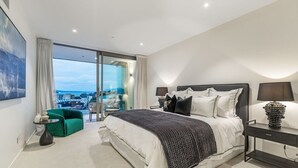 A modern hotel room with a large bed, a green armchair, a glass door leading to a balcony with a city view, and a nightstand with a lamp.