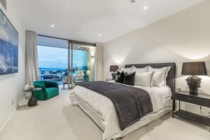 Une chambre d’hôtel moderne avec un grand lit, un fauteuil vert, une porte vitrée donnant sur un balcon avec vue sur la ville, et une table de chevet avec une lampe.