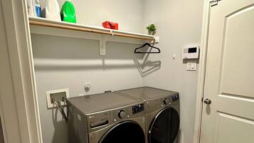 Un cuarto de lavado con dos lavadoras de carga frontal, un estante con productos de limpieza y una puerta.