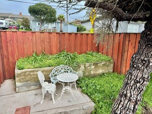 Een terras met een betonnen plantenbak, twee witte metalen stoelen en een kleine ronde tafel.