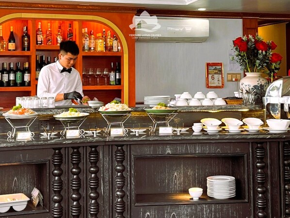 A buffet bar with various dishes, a bar area with bottles, and a decorative vase with red flowers. - Verdure Lotus Classic Cruise on Lan Ha Bay (Tuan Chau)