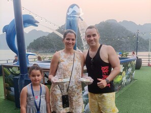 A group of people at a waterfront location with a large dolphin sculpture, a boat, and a mountainous backdrop. - Verdure Lotus Classic Cruise on Lan Ha Bay (Tuan Chau)