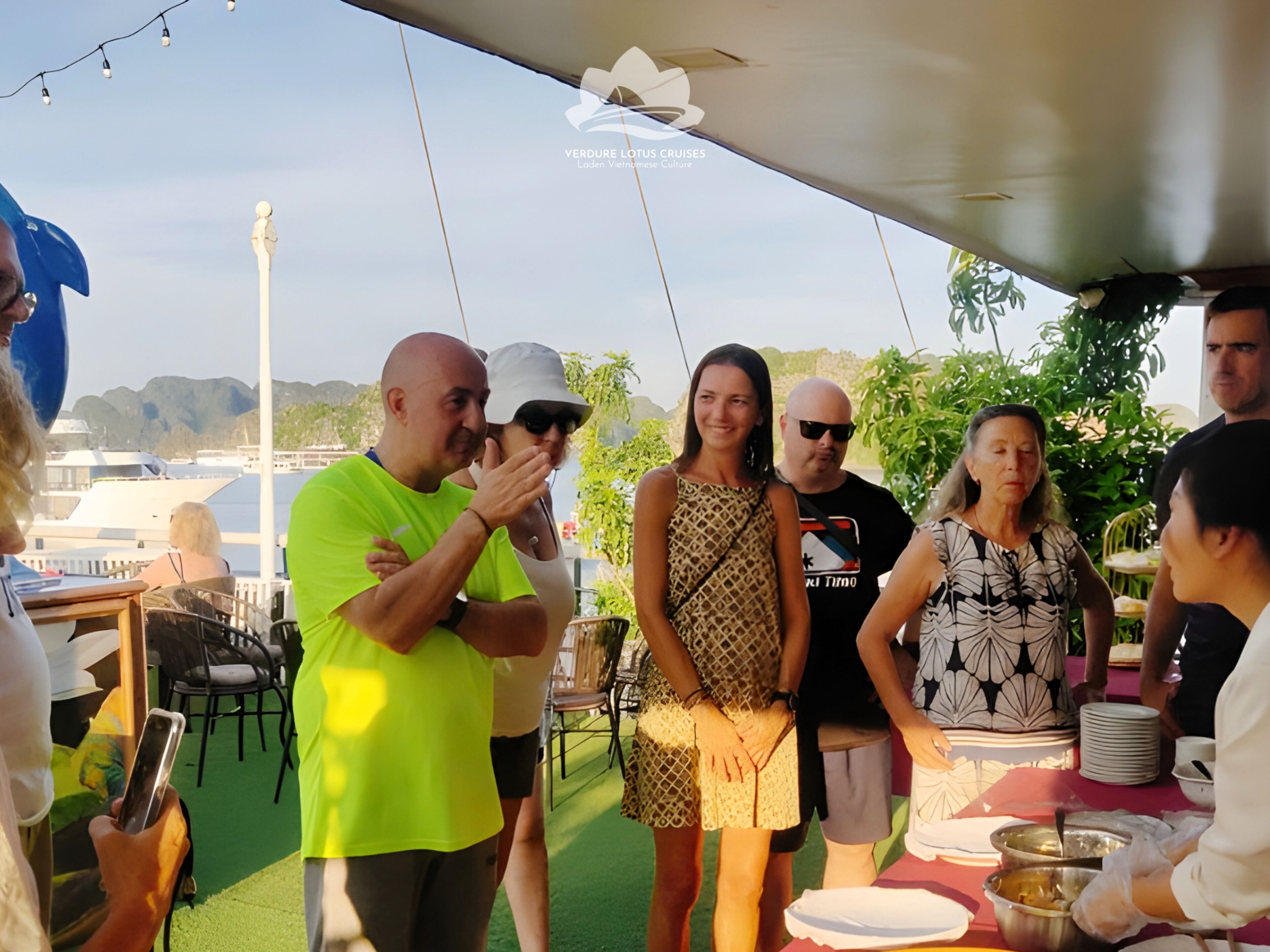A group of people at an outdoor event with a table and chairs, a tent overhead, and a view of boats and greenery in the background.