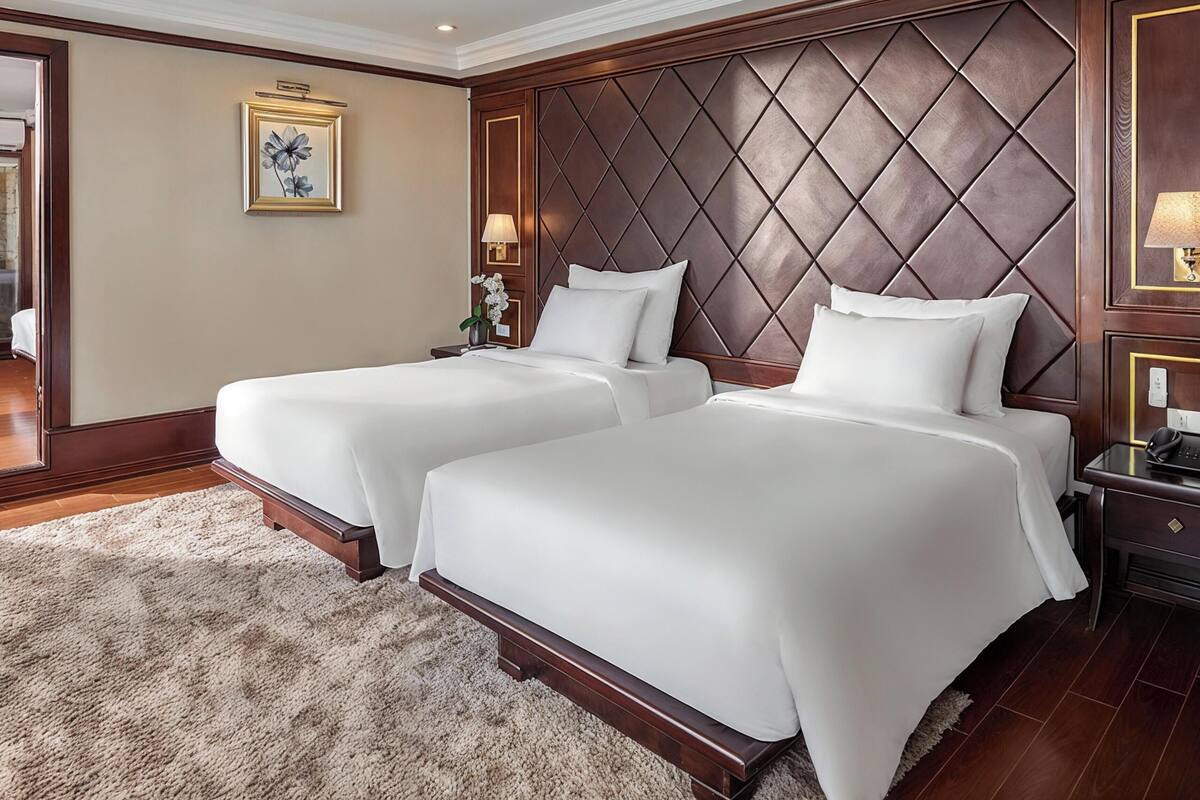 A hotel room with two beds, a large padded headboard, a small bedside table with a lamp, and a framed picture on the wall.
