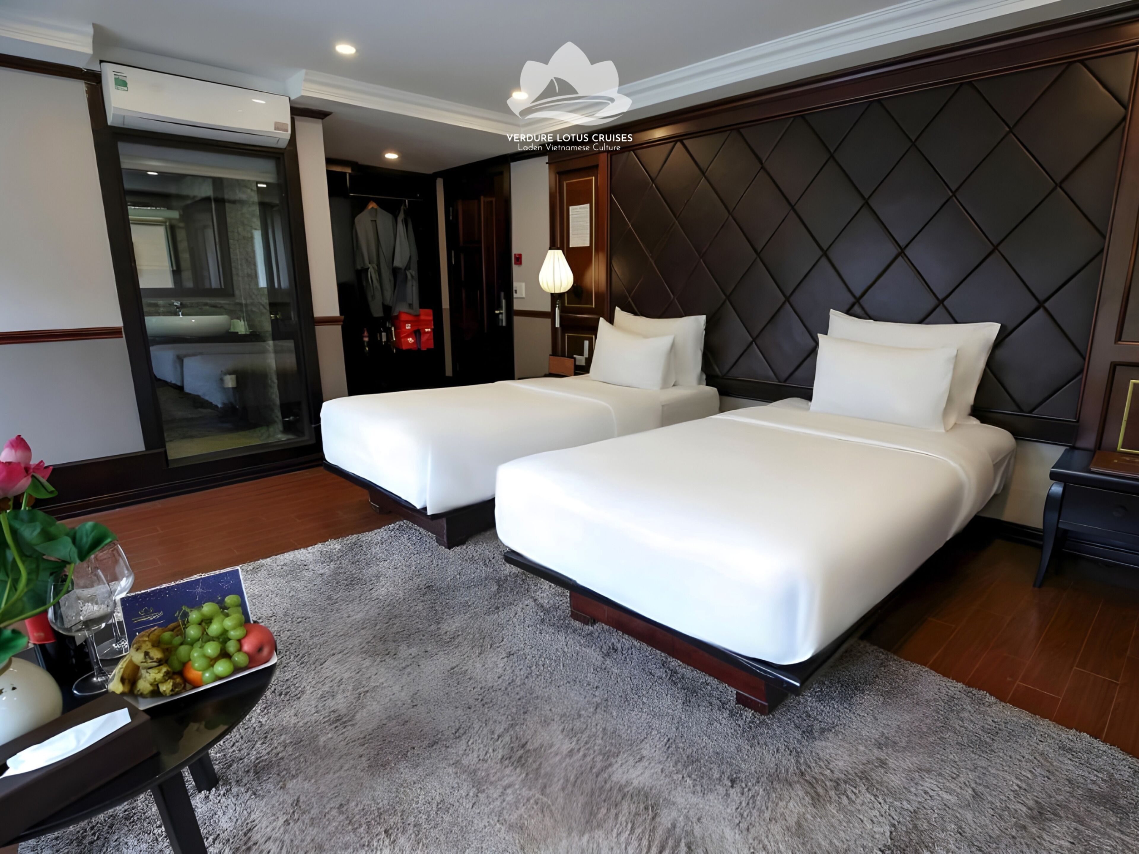A hotel room with two beds, a minibar, a bathroom visible through a glass door, and a small table with fruit.