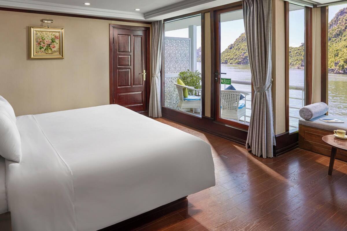 A hotel room with a large bed, a window with a view of a body of water, a door, and a small table with a cup.