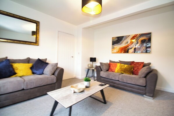 A modern living room with two grey sofas, colorful cushions, a coffee table, a painting on the wall, and a mirror above the sofa.