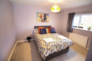 A bedroom with a bed, a nightstand, a lamp, a window with curtains, and a painting on the wall.