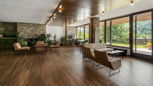 A spacious room with a stone fireplace, wooden flooring, and large windows offering a view of greenery.