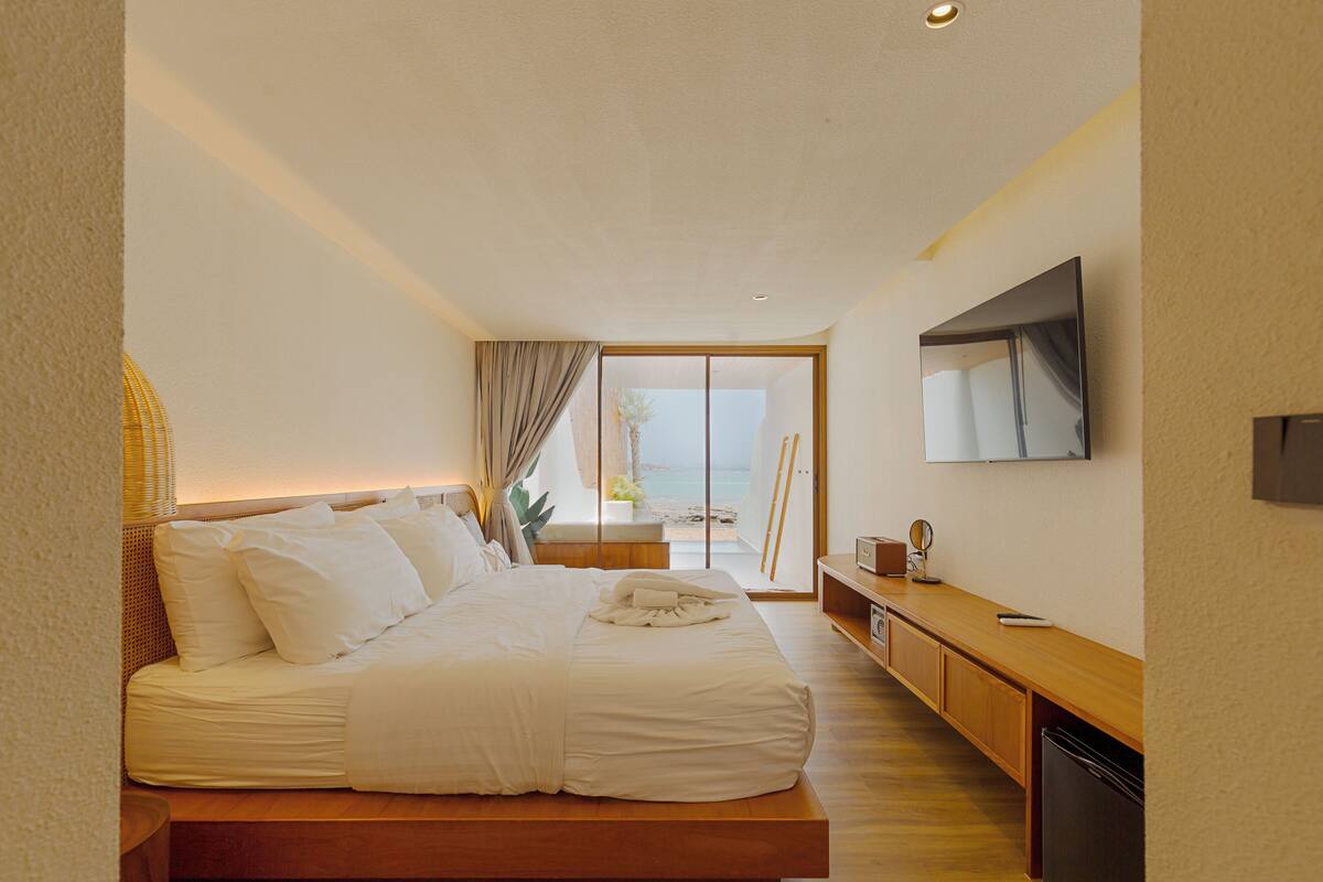 Ein modernes Schlafzimmer mit einem großen Bett, einem an der Wand montierten Fernseher und Blick auf den Strand durch das Fenster.