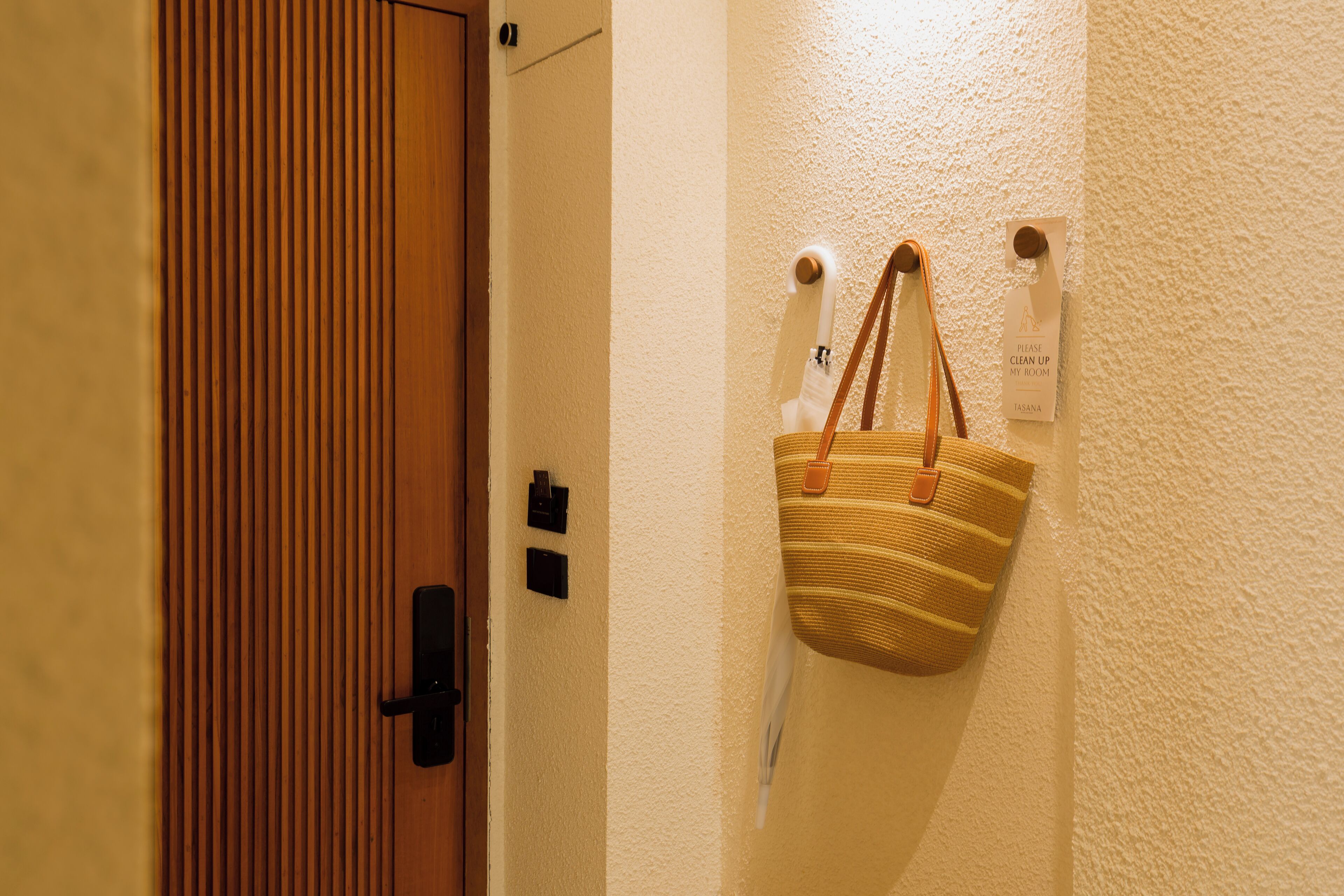 Eine Holztür mit vertikaler Paneeloptik, ein an der Wand befestigter Bag-Halter mit einer gestreiften Tasche und ein Schild mit der Aufschrift 'Bitte räumen Sie mein Zimmer auf'.