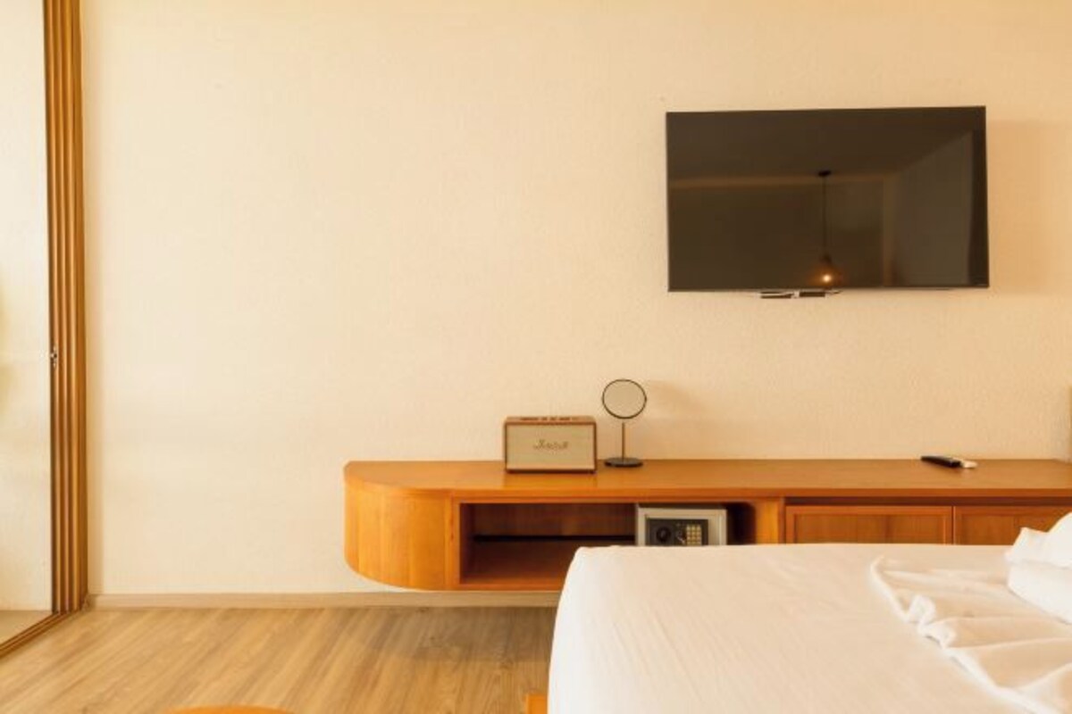 Une chambre d’hôtel avec un lit, une table de chevet en bois, un téléviseur fixé au mur et une petite étagère avec une radio et une horloge.