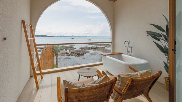 Un balcón con vista al mar, una bañera, sillas de mimbre y una mesita.