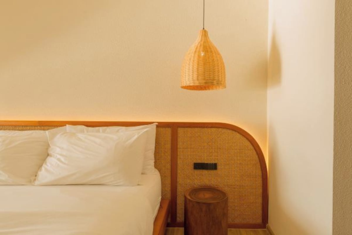 Una cama con ropa de cama blanca, un cabecero tejido y una mesita de noche de madera con un objeto redondo de madera encima.