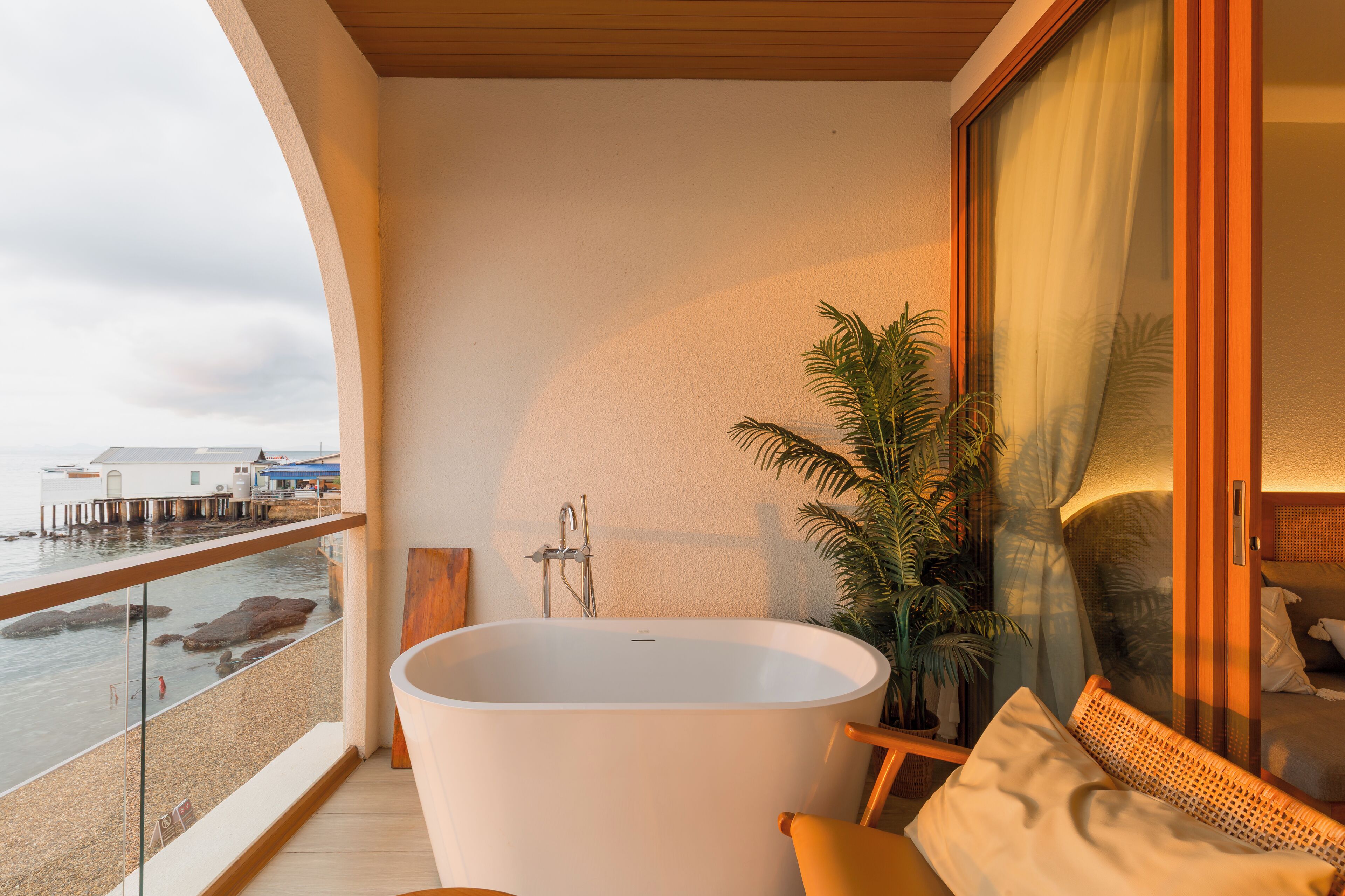 A balcony with a large bathtub, a potted plant, and a view of the sea and buildings.