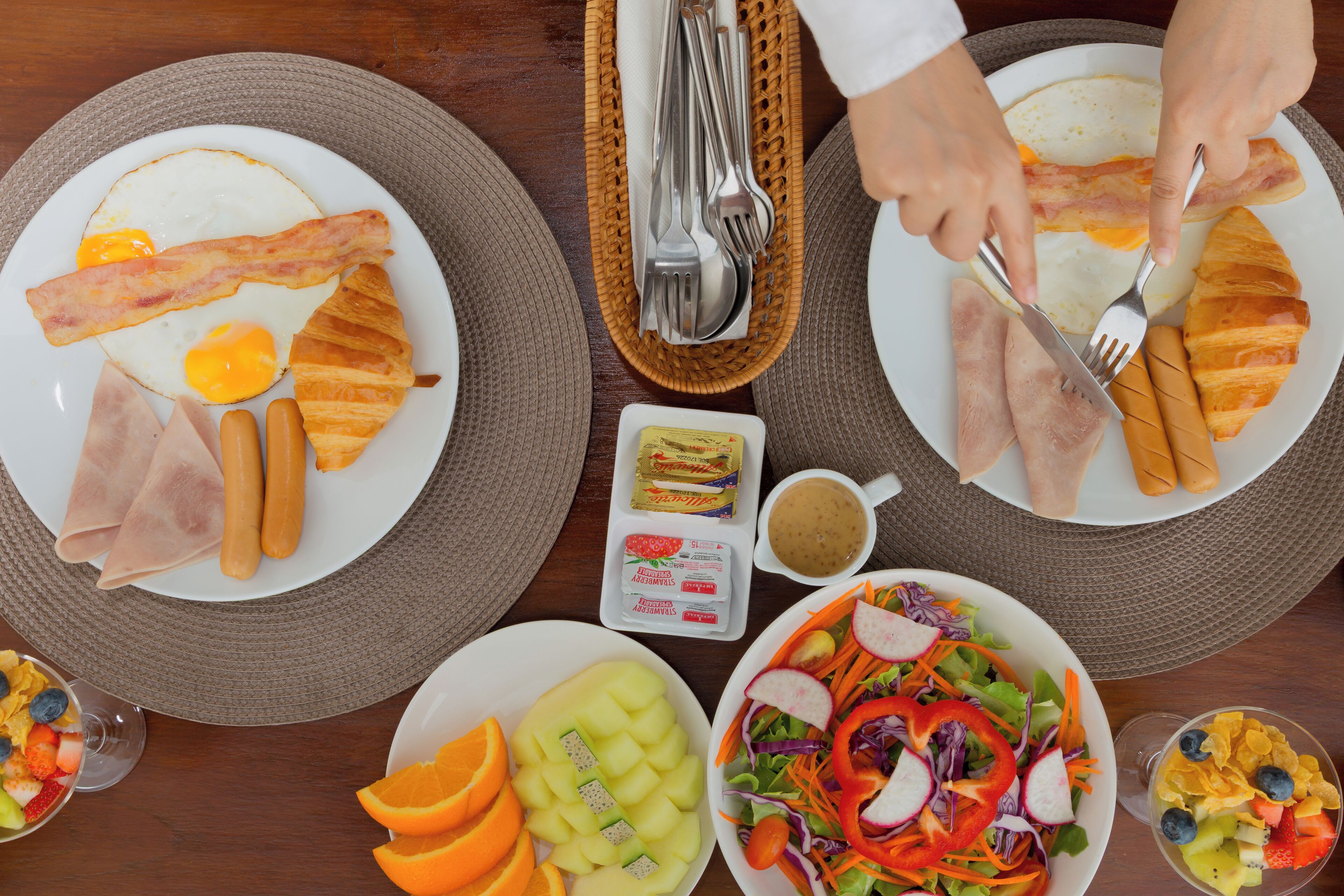 Un desayuno con platos de huevos, tocino y jamón, un tazón de fruta y una ensalada.