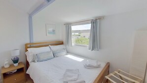 A neatly made bed with white linens and striped pillows, a bedside table with a lamp and a small plant, a window with curtains, and a framed picture on the wall. - G23 Norda, Riviere Towans (Hayle)