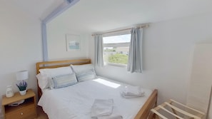 Una cama bien tendida con sábanas blancas y almohadas a rayas, una mesita de noche con una lámpara y una planta pequeña, una ventana con cortinas y un cuadro en la pared.