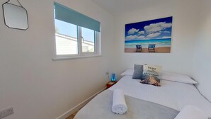 A bedroom with a bed, a window, a painting of a beach scene, and a small table with a lamp.