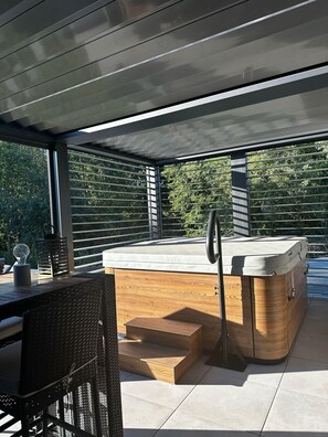 Un espace moderne aménagé à l’extérieur avec un jacuzzi, une terrasse en bois, un coin salon et une pergola.