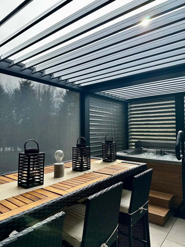Un espace repas extérieur moderne, avec une table et des chaises en bois, des lanternes noires et une vue sur l’extérieur grâce à un plafond à lattes.