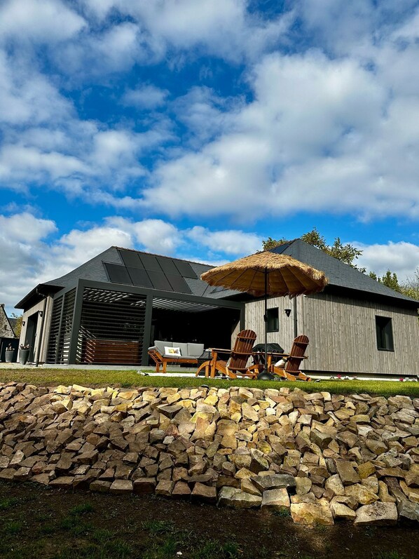 Une maison moderne avec un mur en pierre, un patio couvert avec des chaises et un grand parasol.