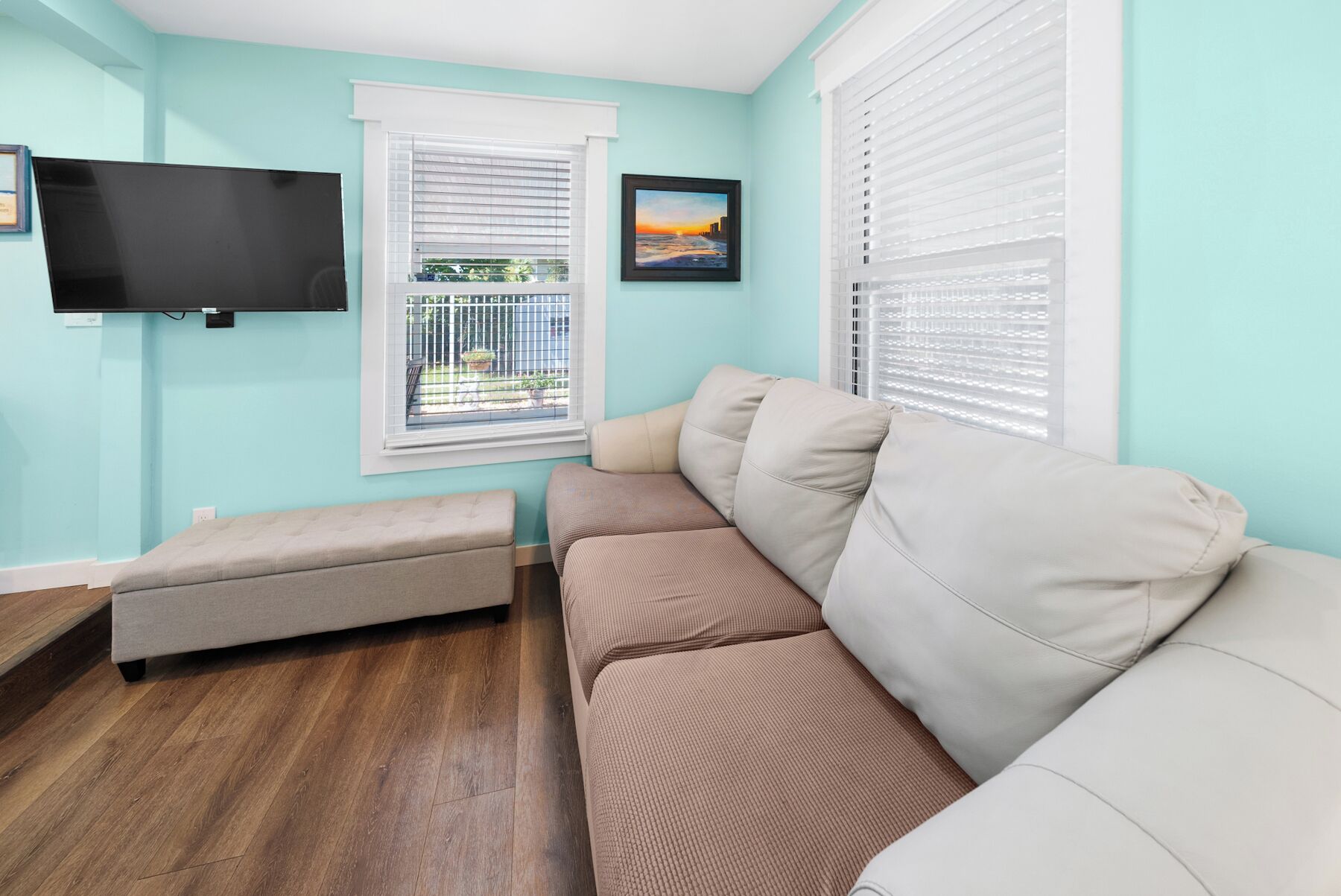A living room with a beige sofa, a small grey sofa, a TV mounted on the wall, and a window with blinds.