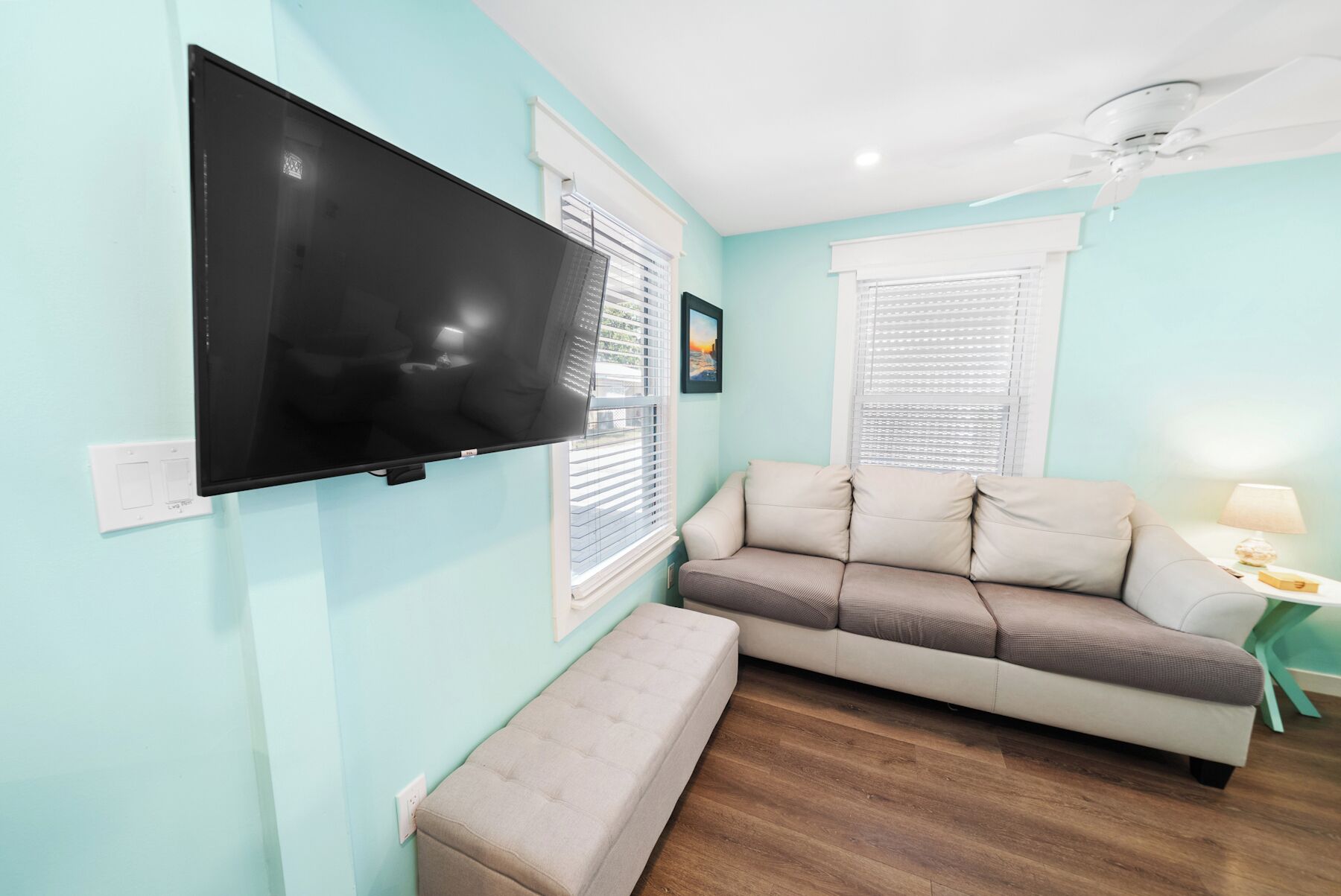 Ein Wohnzimmer mit Flachbildfernseher an der Wand, einem beigefarbenen Sofa, einem kleinen Tisch mit einer Lampe und einem Deckenventilator.
