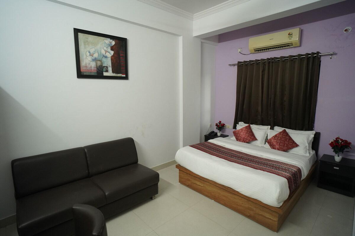 A bedroom with a bed, a dark sofa, a nightstand, an air conditioner, and a framed painting on the wall.