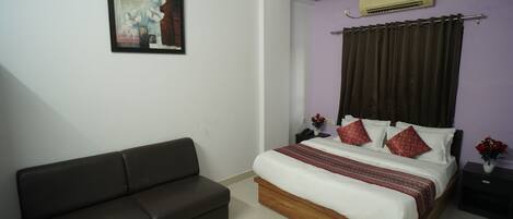 Un dormitorio con cama, un sofá oscuro, una mesita de noche, un aire acondicionado y un cuadro enmarcado en la pared.