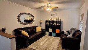Una sala de estar con un sofá de cuero marrón oscuro, un sillón a juego, un espejo decorativo, un ventilador de techo y un mueble de almacenamiento negro con cajones de colores.