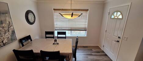 A dining room with a table, chairs, a clock, and a window with blinds.